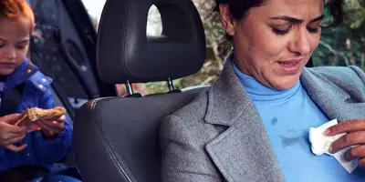 Een vrouw zit op de voorstoel van een auto haar blouse schoon te maken; een meisje eet een boterham op de achterbank.