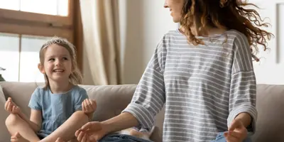 Una donna con maglia a righe e una bambina meditano sorridenti sul divano del salotto con le gambe incrociate.