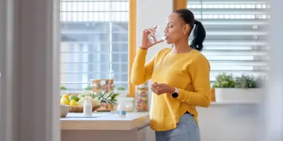 Una donna con coda di cavallo e maglione giallo beve un bicchiere d'acqua in piedi in una cucina.