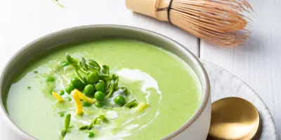 Grüne Matcha-Erbsensuppe garniert mit Erbsen und Zitronenschale in einer Schüssel. Daneben ein goldener Löffel und ein Matcha Besen.