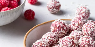 Mehrere pinke Himbeer-Energy-Balls mit Kokosraspeln in einer hellen Schüssel. Im Hintergrund steht eine Schüssel mit Himbeeren.